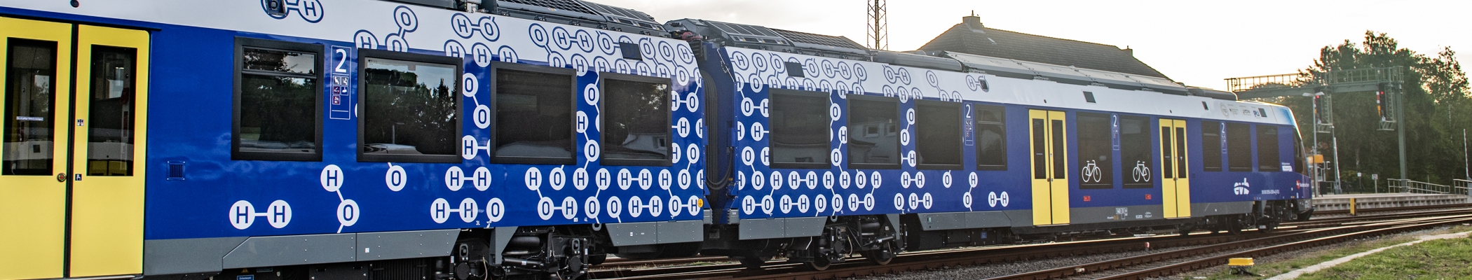 Blau-weiß-gelbes Wasserstofffahrzeug für den SPNV in Niedersachsen in extremer Seitenansicht mit Wasserstoff-Formel-Symbolen, in Fahrtrichtung nach rechts - Fotografin: Sabrina Adeline Nagel